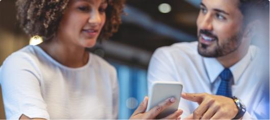 Businesswoman and man smiling while looking at smartphone, surrounded by paperwork and a laptop.
