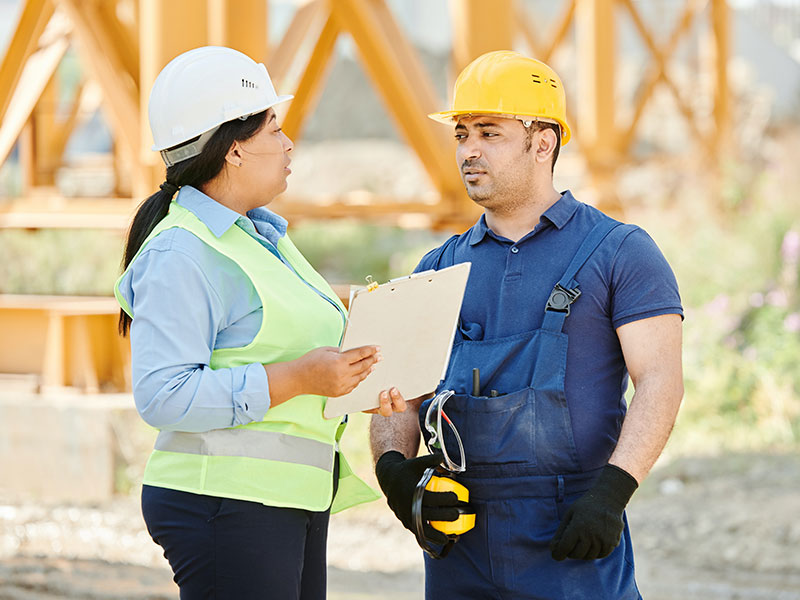 A woman in a white hard hat and green vest holding a clipboard talking to a man in a yellow hard hat and blue shirt.