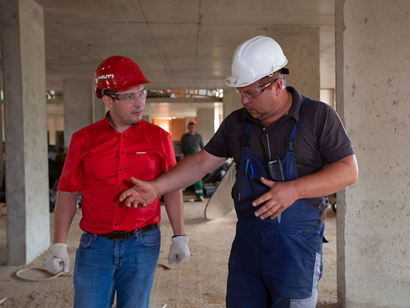 Man in a red shirt and red construction helmet talks to a man with blue overalls and white hat at a construction site.