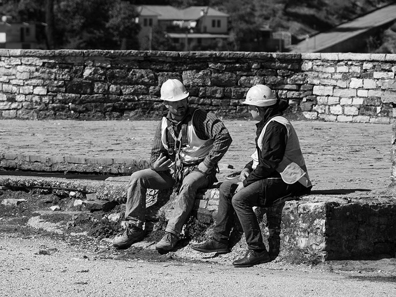 Two construction workers wearing hard hats and vests sitting on a stone wall, grayscale