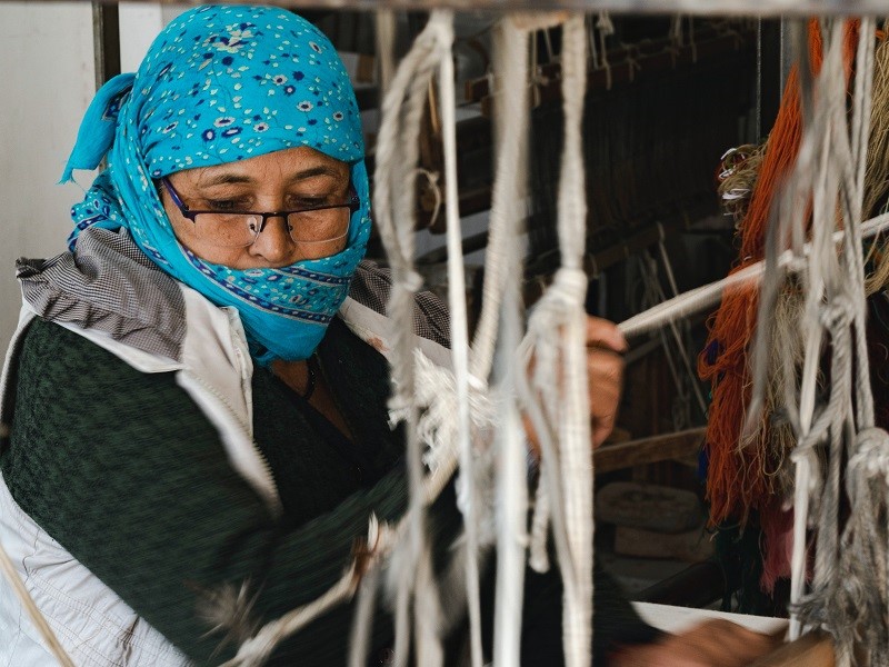 A woman in a blue headscarf wearing a mask and using a hand loom to weave