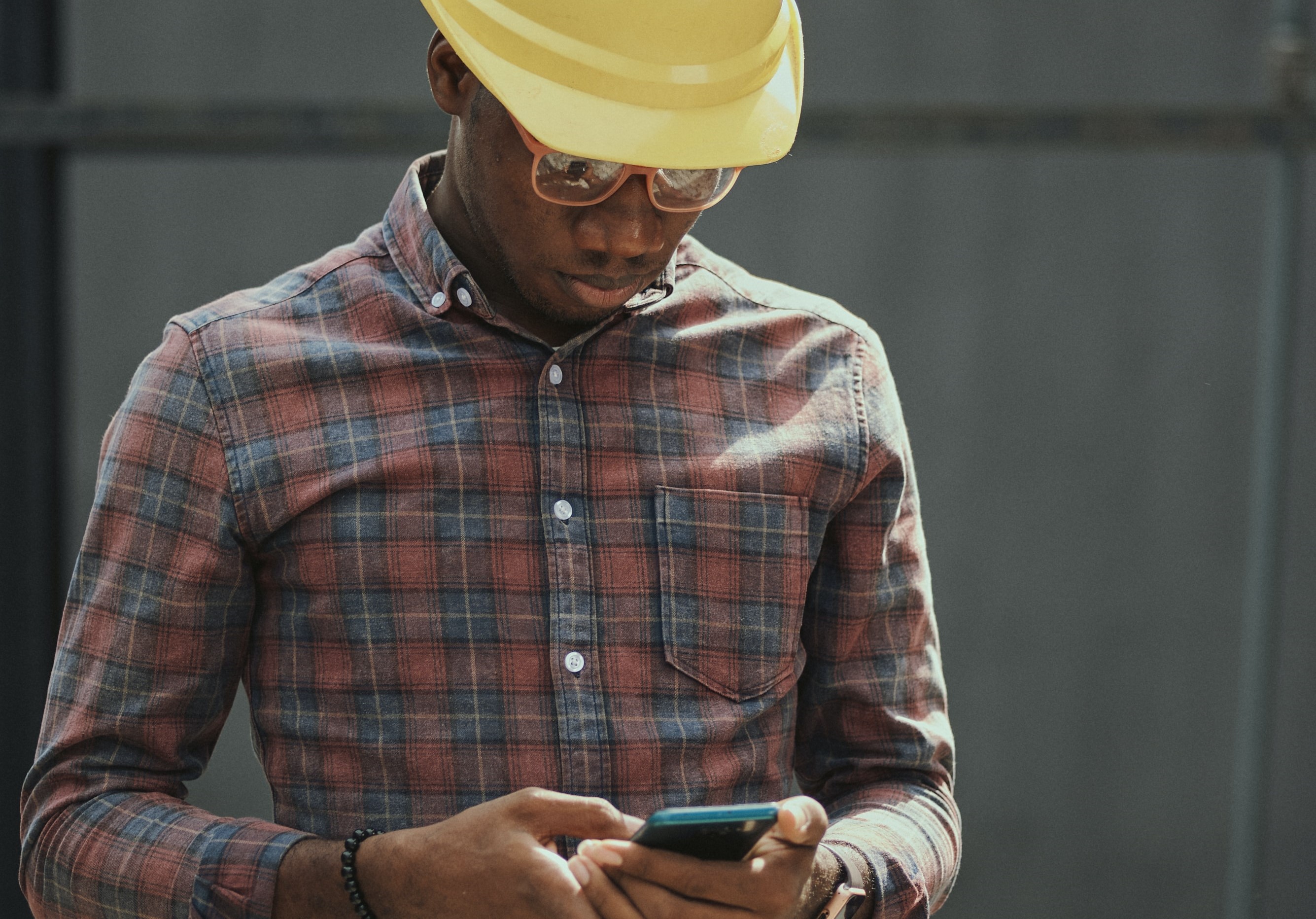 Construction worker in red, blue and white plaid dress shirt wearing yellow hat holding black smartphone