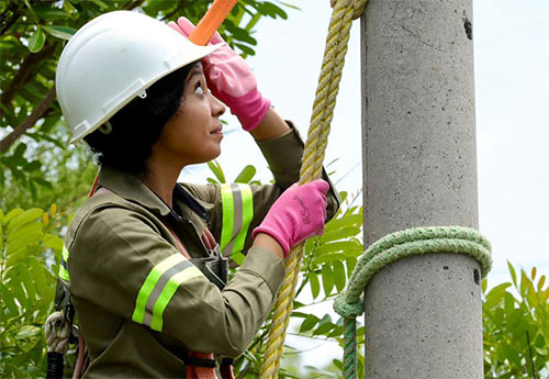 A woman in uniform and a hard hat uses a rope to anchor herself to a pole. Tree tops are in the background.
