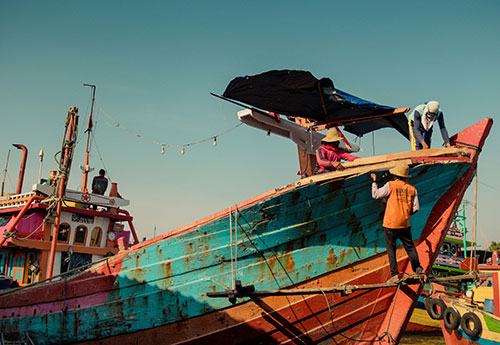 A fishing vessel undergoing maintenance in Juwana, Pati Regency, Central Java, Indonesia.  Two people are on board, and one person stands on a platform below the ship. Photo by Alfi Hilman for Unsplash.