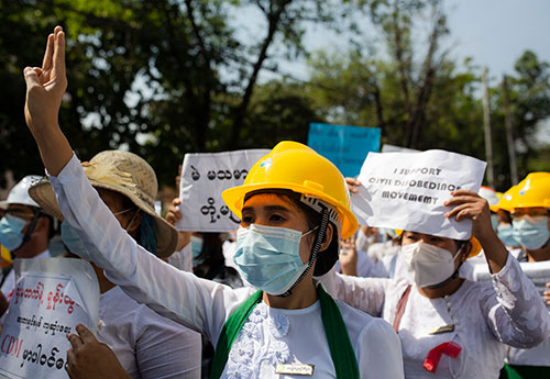 A women wearing a hard hat and surgical mask stands among a crowd of workers with her hand in the air. Many of the workers are carrying signs.
