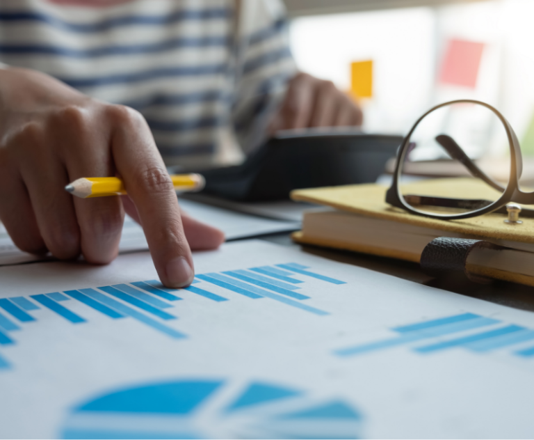 Person analyzing charts with a pencil in hand. Eye glasses and books are on the table.