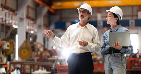 Two workers wearing hard hats stand in an industrial facility, one gesturing while the other holds a laptop.