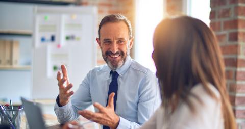 Business professional smiling and gesturing while discussing succession planning with a colleague at a desk. 