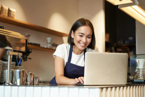 An owner of a coffee shop works on a laptop behind the coffee counter.