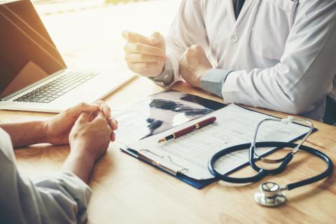 A patient and doctor have a conversation at a desk in a doctor's office.
