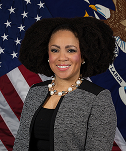 Assistant Secretary Taryn Williams in a black and white suit and pearls. The DOL flag is in the background.