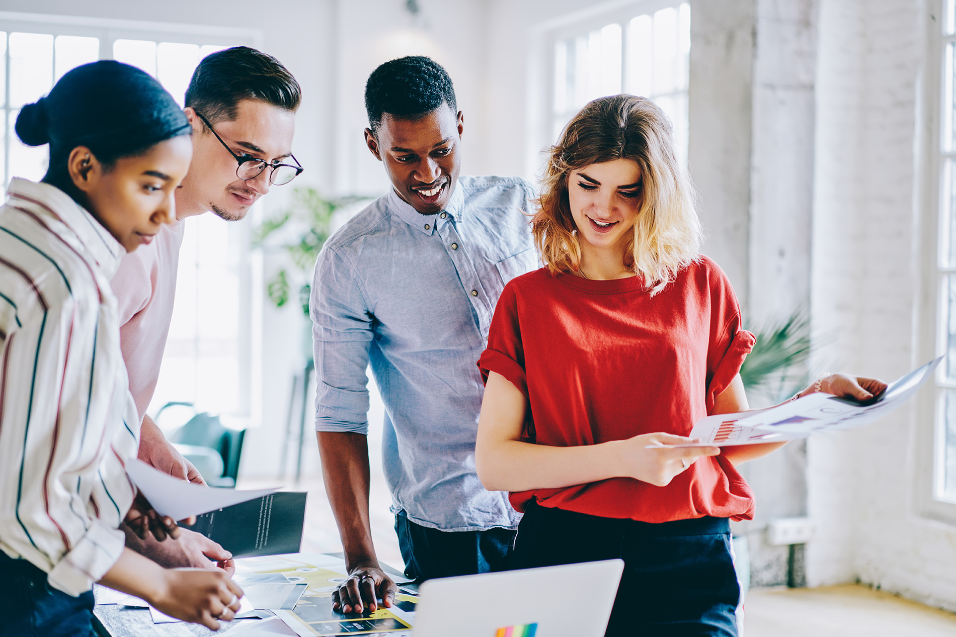 Four coworkers standing around a table reviewing printed documents and a laptop in a bright office.