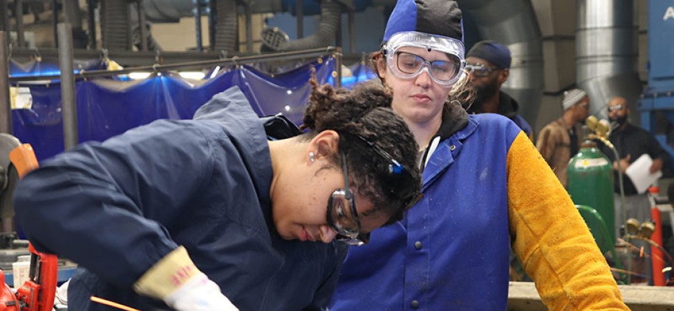 Person in blue long sleeve shirt and blue vest using a welder while being supervised. Safety, guidance, supervision