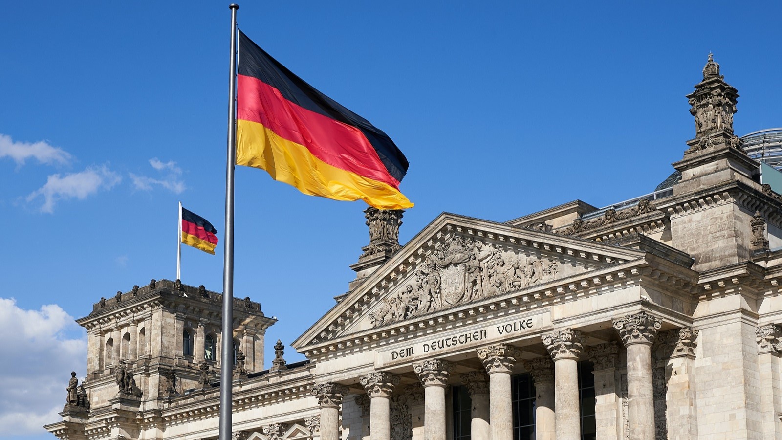 Black,red, and yellow German flag on a pole outside of large stone building with columns