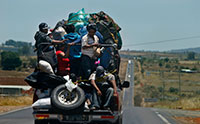 A group of people is packed onto the bag of a truck with bags and equipment driving down a hilly road.