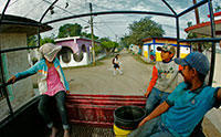 A man, woman, and child ride in the back of a pick-up truck with a bucket. A young girl walks behind them on the road.