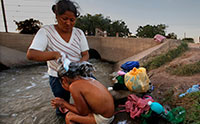 A woman stands and washes a young girls hair on the banks of a muddy canal.