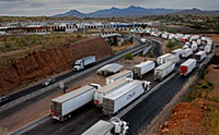 Tractor trailer trucks lined up on a highway with a cluster of buildings and mountains in the background.
