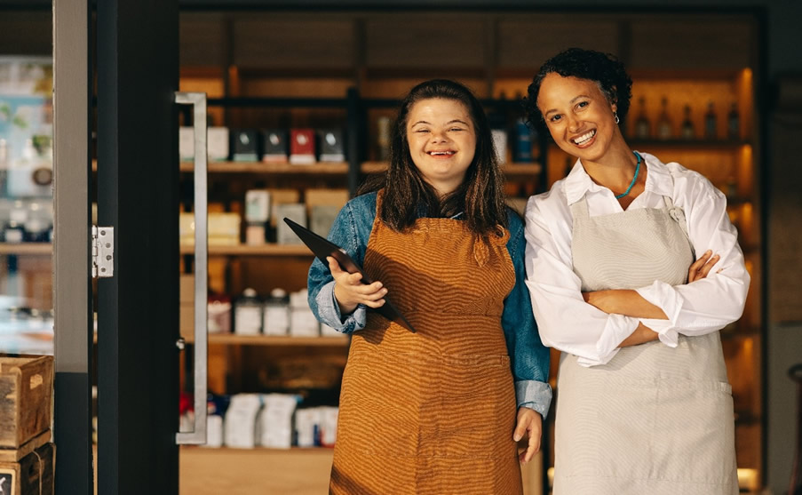 On the left is a smiling young woman wearing a blue denim shirt with an orange apron standing with a more mature woman wearing a white blouse and cream apron to her right, who is also smiling. The young woman has a disability and is confident and happy working at her retail job. 