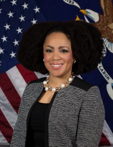 Pictured is a Black woman with shoulder length natural hair smiling at the camera in front of the American and U.S. Department of Labor flags