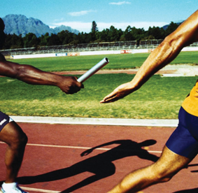Two men running on a track in a relay race.  One person is passing the baton to the other.