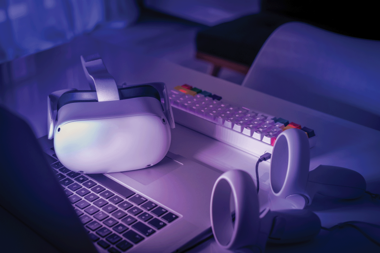 Laptop, virtual reality headset, multimedia keyboard and air joysticks on a desk.