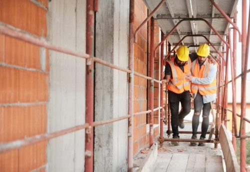 Two workers wearing hard hats and safety vests walking on scaffolding at a construction site, one worker assisting the other.New Videos! How RETAIN’s Stay-at-Work/Return-to-Work Program Changes Lives