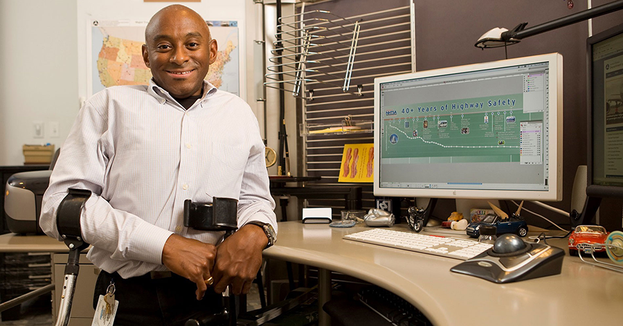 A young person using forearm crutches stands and smiles at a desk with a computer.