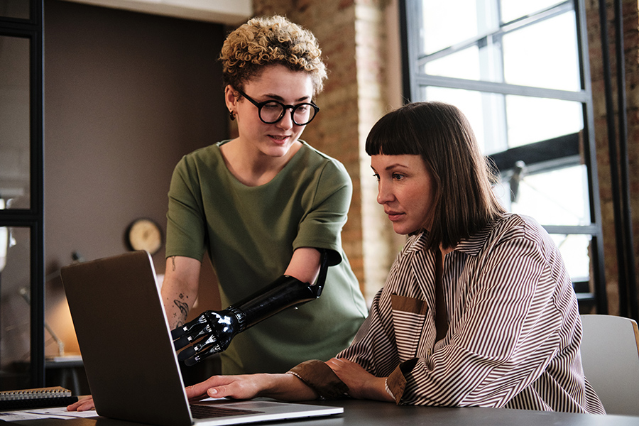 Two people looking at a computer. One person has short curly blond hair with glasses, a prosthetic arm, and an olive green shirt. The other has straight shoulder-length brown hair with bangs and a brown and white striped blouse.
