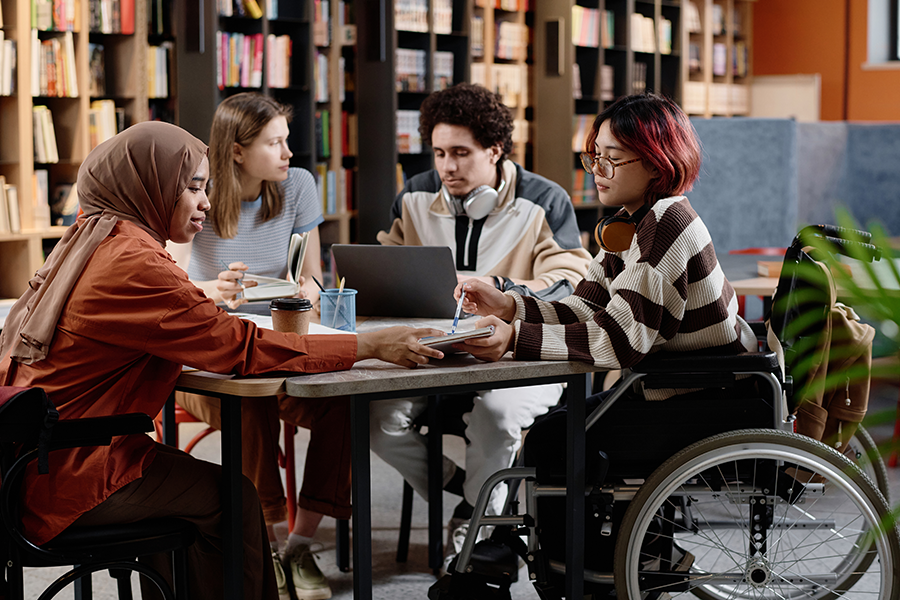 Four young adults, including one using a wheelchair, sit and collaborate at a library table with a computer, notebook, and coffee. 
