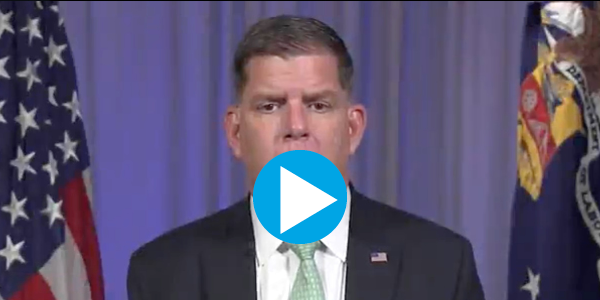 A still frame from a video featuring Secretary Walsh sitting in front of a blue curtain in between an American flag and a flag bearing the Labor Department seal. A play button appears in the foreground.  