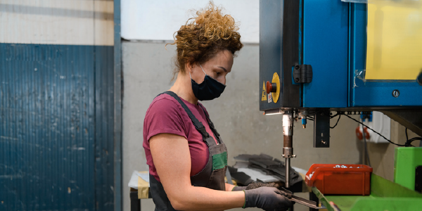 A woman wearing a mask and protective apron works with a metal drill in a workshop.  