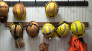 Photo shows eight mining helmets hanging on a wall along with protective gloves and a reflective safety vest. 