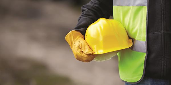A close-up shot shows the torso of a worker in a safety vest and protective gloves holding a hard hat. 