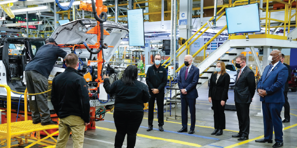 President Biden and Secretary Walsh stand on the floor of an auto manufacturing factory with GMs Mary Barra and UAWs Ray Curry.