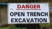 Sign on a fence reads: Danger, open trench excavation.