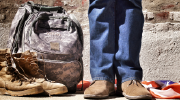 Close-up shot of a person from the knees down, wearing jeans and civilian shoes, standing next to military boots and a backpack in front of a U.S. flag.
