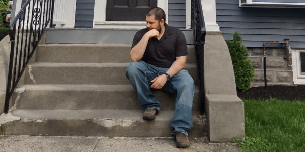 A man sits on the stairs in front of a house, gazing into the distance.