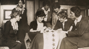 Black and white photo: National Woman's Party activists watch Alice Paul sew a star onto a flag, representing another state's ratification of the 19th Amendment.