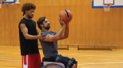 A personal trainer helps a man in a wheelchair perfect his form while shooting baskets. 