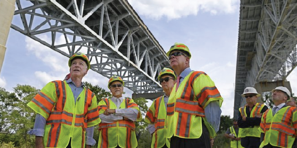 Secretary Walsh stands with a group of men, all in safety vests and hard hats, beneath a bridge undergoing construction. 