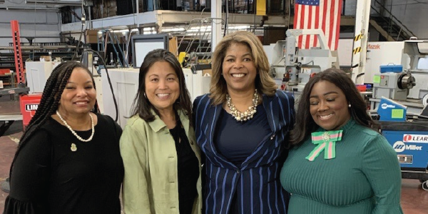 Deputy Secretary Julie Su stands with three professionally dressed women in a factory. All four women are smiling at the camera.