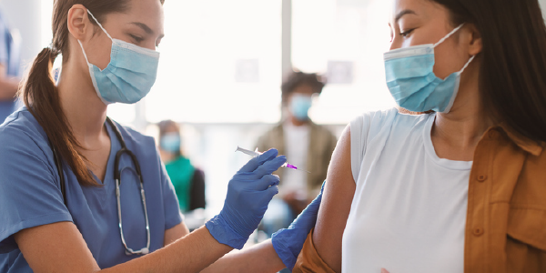 A woman in scrubs, wearing a mask and medical gloves, administers a vaccine to another woman wearing a surgical mask.