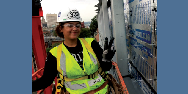 A confident young woman smiling and giving the peace sign while standing on an aerial lift in her uniform and safety gear. City buildings are visible behind her.