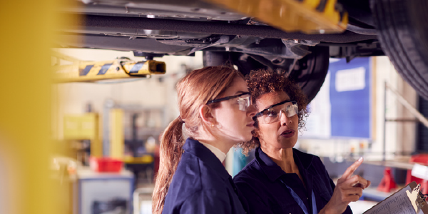Two women in mechanics’ coveralls and safety glasses examine a car on a lift.
