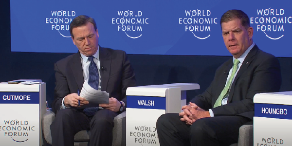 Secretary Walsh speaks as part of a panel. He is seated between a man in a suit, whose nameplate reads 'Cutmore,' and an unseen man whose nameplate reads 'Houngbo.' World Economic Forum icons cover the wall behind them.