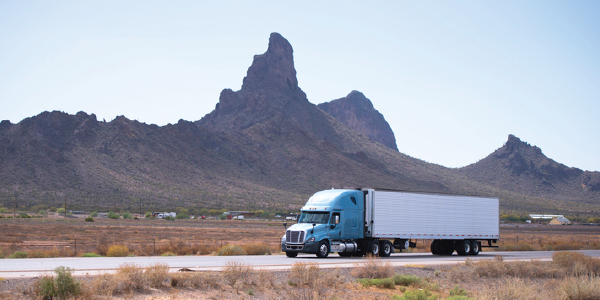 An 18-wheeler truck moves down a highway with arid plants in the foreground and dramatic rock formations rising in the background.