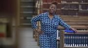 Riquita Quarterman, wearing a blue dress, stands in a library next to a shelf of reference materials.