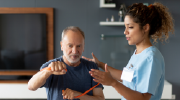 A young woman in scrubs guides an older man through physical therapy exercises using a resistance band.