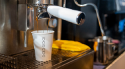 Close-up shot of a commercial espresso machine in use. Coffee drips into a white paper cup.